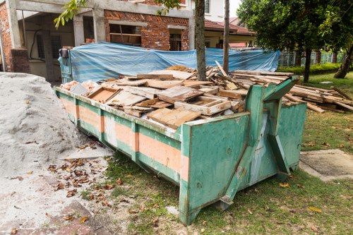 A large open-top green skip filled with a pile of wooden debris, including planks, panels, and construction waste, situated on a grassy area beside a sidewalk in a residential or semi-urban environment. The skip shows signs of use with some rust and dirt on its surface. Behind it, there is a partially constructed building with exposed brickwork and scaffolding, along with a blue tarp covering part of the construction site. To the right, there is a tree with green foliage, and nearby, other trees and bushes are visible. The scene appears to be outdoors during daylight, with natural lighting illuminating the area, illustrating a typical rubbish removal setting that Commercial Waste Harlow might service in the Harlow region.