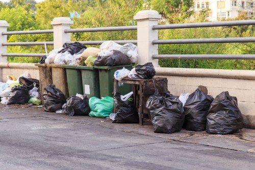 A collection of black, green, and white plastic rubbish bags filled with waste is stacked along the edge of a paved street or pathway, positioned under a metal railing with horizontal bars supported by concrete posts. Some of the bags are leaning against the green waste bins, which are partially visible and appear to be designed for waste collection. The bags vary in size and are tightly sealed, indicating they are ready for disposal. In the background, there are trees and possibly residential or office buildings, suggesting an urban or suburban environment typical of a town in the UK. The scene is illuminated by natural daylight, casting soft shadows and highlighting the mixture of textures and colors of the rubbish bags against the smooth pavement surface. This image is relevant to local rubbish removal services, such as those offered by Commercial Waste Harlow, serving areas like Harlow and surrounding postcode districts, and reflects typical street-level waste collection scenarios commonly managed by professional waste management companies.