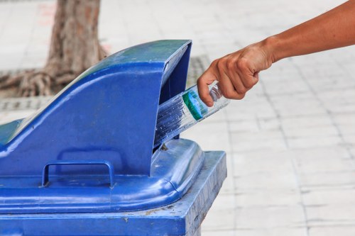 A person's hand is shown placing a clear plastic water bottle into a blue recycling bin, which is situated on a paved pedestrian area. The water bottle appears to be partially squeezed, with a green and white label visible around its middle. The bin has a rounded lid with an opening specifically designed for bottles, and the surface of the bin shows slight weathering and dirt marks typical of outdoor waste containers. In the background, a blurred tree trunk and the pavement are visible, indicating an outdoor urban setting likely within Harlow or its surrounding areas. The image emphasizes proper waste disposal practices relevant to rubbish removal services offered by Commercial Waste Harlow in Hertfordshire, supporting responsible recycling and waste management efforts in the local community.