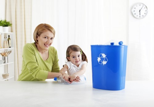 A woman with short, light brown hair wearing a lime green top sits at a white kitchen counter alongside a young girl with dark hair in pigtails, dressed in a white shirt. They are smiling as they both position a clear plastic waste bag onto an indoor surface. To the right of the image, there is a large, bright blue recycling bin with a white recycling symbol printed on its front, situated in a clean, well-lit kitchen environment with a white wall, a round wall clock displaying approximately 1:00, and decorative items on a shelf in the background. The scene appears to be related to waste management or recycling activities, illustrating domestic rubbish disposal or collection, which aligns with services offered by Commercial Waste Harlow for rubbish removal in the local area. The setting emphasizes a tidy, organized approach to recycling and waste management in a home environment, subtly supporting local SEO signals for rubbish removal services in Harlow.