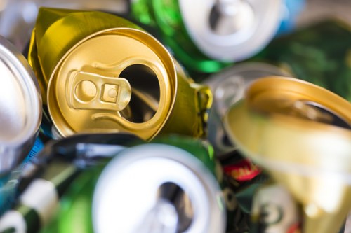 A close-up image showcasing a collection of crushed aluminum beverage cans, predominantly in gold, silver, and green colors, with some cans displaying pull tabs and others with their tops removed. The cans are piled together on a surface, with textures reflecting their metallic finishes, some appearing glossy while others are matte. The surrounding environment suggests an indoor or warehouse setting, with a focus on waste materials related to rubbish removal services. The cans are stacked and slightly overlapping, illustrating typical recyclable waste items that a commercial waste removal company like Commercial Waste Harlow might handle. The image emphasizes the importance of proper disposal and recycling of aluminum cans within the context of rubbish management in Harlow or nearby areas of Essex, subtly supporting local SEO relevance through the focus on common recyclable packaging materials.