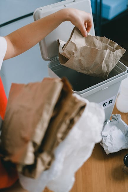 A large pile of mixed waste, including cardboard boxes, plastic bags, discarded paper, and miscellaneous packaging, is overflowing from several open rubbish bins on a paved urban street. The backgroun